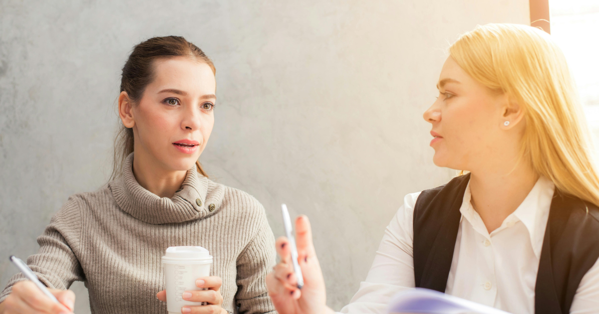 Two women discussing toxic stress and healing. One holds a coffee cup while taking notes, the other gestures with a pen.